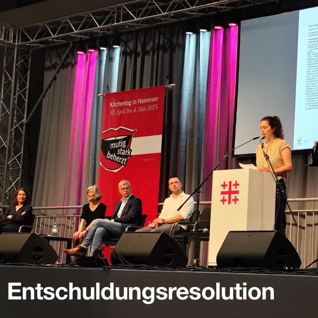 Bild zeigt fünf Menschen auf einer Bühne des evangelischen Kirchentages 2025 in Hannover. Eine Person davon steht hinter einem Pult und spricht in zwei Mikrofone. Text: Entschuldungsresolution.