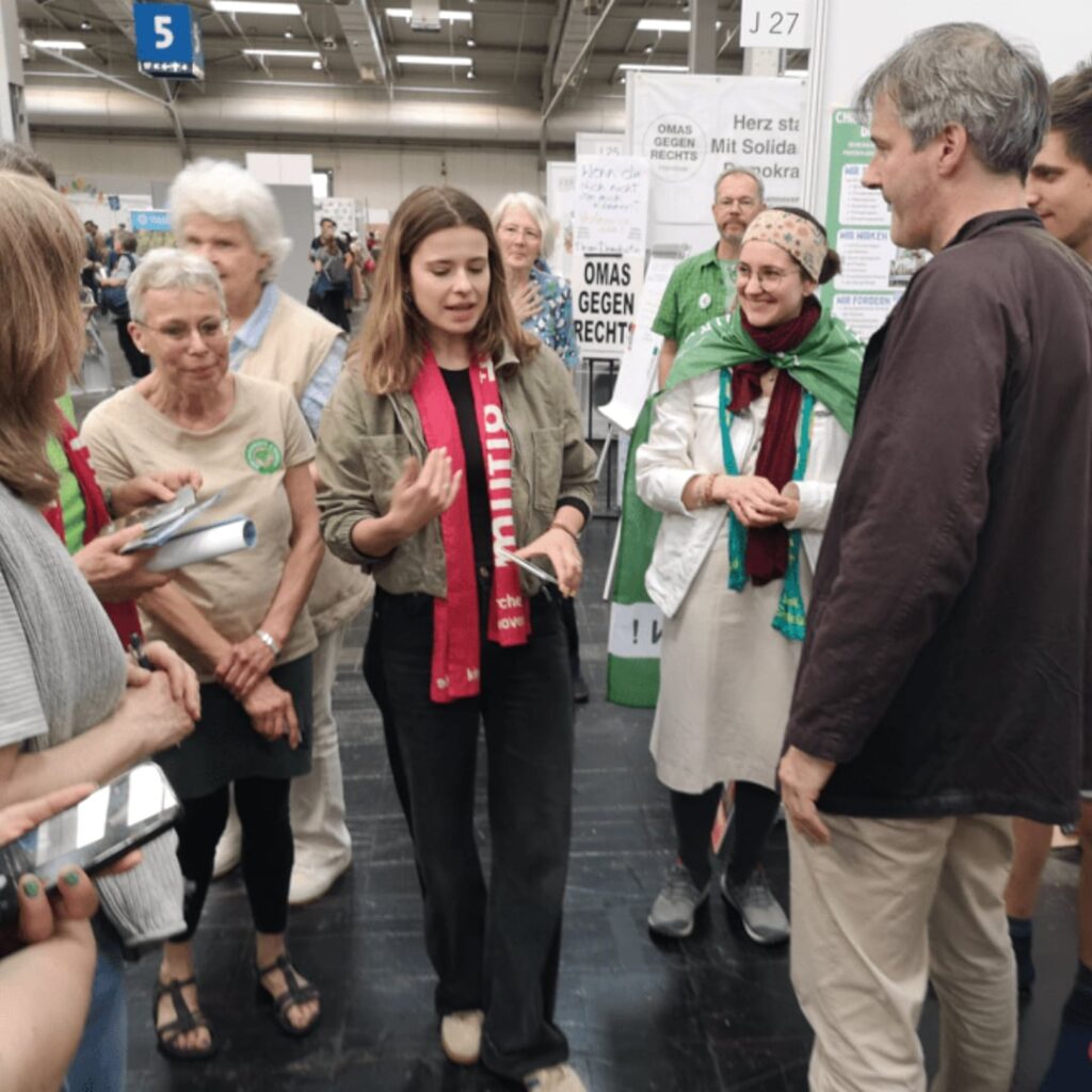 Luisa Neubauer in der Mette des Bildes, diskutiert mit umstehenden Personen am Stand der C4F auf dem Kirchentag 2025 in Hannover.