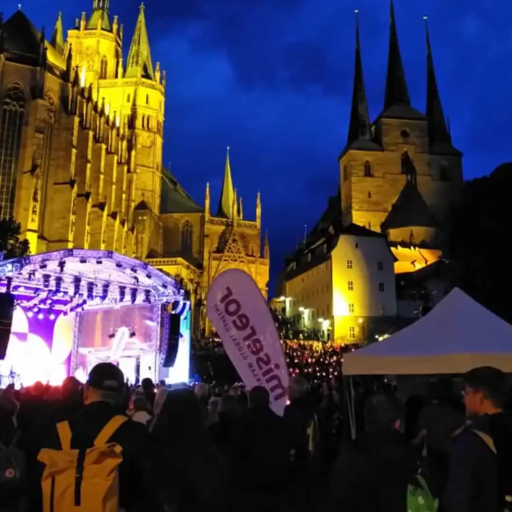 Das Bild zeigt eine abendliche Stimmung vom Katholik*innentag in Erfurt 2024. Im Hintergrund der Dom und im Vordergrund eine große Menschenmenge mit Lichtern.