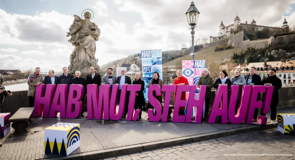 Das Bild zeigt vor der Kulisse Würzburgs im Hintergrund, Menschen, die in einer Reihe auf einer Brücke stehen. Vor den menschen stehen meterhohe Buchstaben, die das Motto des Kirchentages bilden: Hab Mut, steh auf!